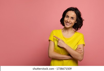 Pointing. Close-up photo of a happy woman in yellow t-shirt, who is looking in the camera and pointing to the left with her  left index finger.