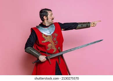 Pointing At Enemy. Comic Portrait Of Medieval Warrior Or Knight Wearing Wearing Armor Clothing Holding Big Sword Isolated Over Pink Background. Comparison Of Eras, History, Funny Meme Emotions