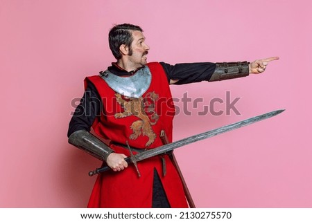 Pointing at enemy. Comic portrait of medieval warrior or knight wearing wearing armor clothing holding big sword isolated over pink background. Comparison of eras, history, funny meme emotions