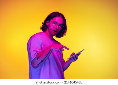 Pointing At Phone. Portrait Of Young Man With Long Hair Using Digital Gadget Isolated On Orange Background In Purple Neon Light. Concept Of Emotions, Aspiration, Sales. Copy Space For Ad.