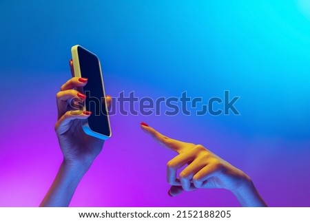 Pointing at phone screen. Closeup female hands holding gadget, smartphone isolated on gradient blue and purple background in neon. Concept of mobile lifestyle, digital technology, social gathering.