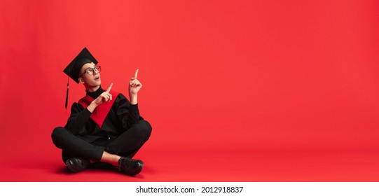 Pointing Upwards. One Young Smiling Student In Mantle And Mortarboard Sitting On Floor Posing On Red Studio Background. Education, Graduation And Student Life Concept.