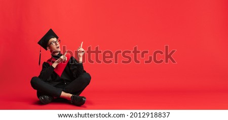 Pointing upwards. One young smiling student in mantle and mortarboard sitting on floor posing on red studio background. Education, graduation and student life concept.