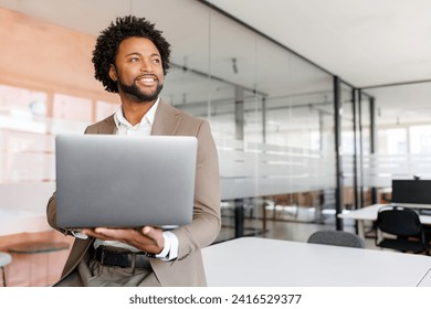 Poised African American businessman in a taupe suit holds his laptop and looks off into the distance, reflecting confidence and vision in a modern office setting standing in bright and open workspace - Powered by Shutterstock