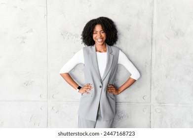 A poised African-American businesswoman stands with hands on her hips, wearing a stylish grey sleeveless vest and trousers, radiating confidence and professionalism against a textured grey background. - Powered by Shutterstock