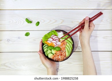 Poke Bowl With Salmon, Rice And Vegetables. Women's Hands Hold Chopsticks And A Bowl With Hawaiian Ahi, A Diet Meal With Fish. Top View.