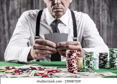 Poker player. Close-up of serious senior man in shirt and suspenders sitting at the poker table and holding cards  with money and  gambling chips laying all around him