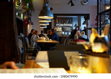 Poland, Gdańsk, March 2020. The Interior Of The Restaurant. In The Foreground, A Blurry Table With A Menu And A Glass. In The Background, People Eating At The Table, Hanging Lamps, Restaurant Logo. 