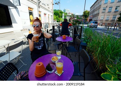 Poland, Warsaw, July 2021. Pub Garden. Colorful Tables With Beer And Snacks, A Woman In Sunglasses Is Holding A Beer In Her Hand, A Sitting Man In The Background. Plants And Building Around.