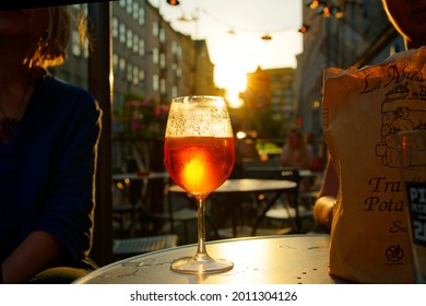 Poland, Warsaw, July 2021. Sunset In The Restaurant Garden. On The Table A Glass Of Aperol Spritz With The Sun Breaking Through. In The Background, Buildings, Pub Lamps, The Sky. A Hot Summer Evening.