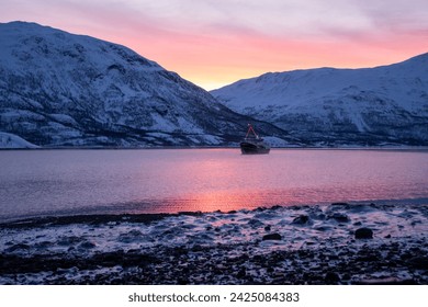 Polar night with small expedition boat anchored in a small bay.
Pink sky and snow covered mountains in the background and frozen sea water icen on the beach in the foreground.
