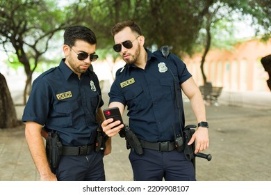 Police Agents With Sunglasses Looking At A Text Message Or A Crime Report On The Smartphone While Chasing A Criminal With GPS