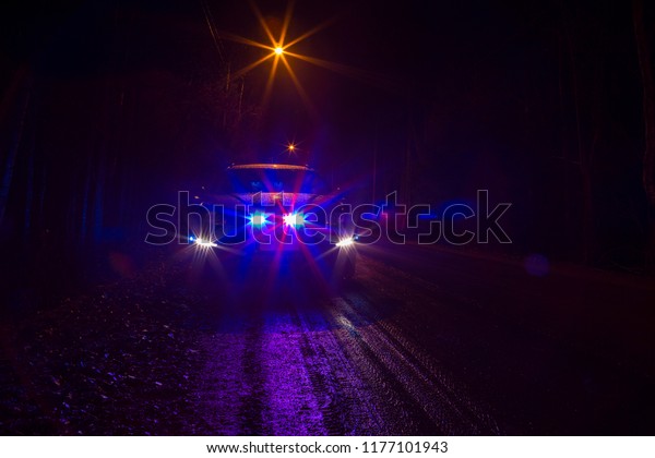 Police car on a dark road flashing blue and red\
lights. Rainy and dark weather. Image has a noise and flare effect\
applied.