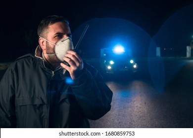 Police Man With Surgical Mask Talking With Walkie Talkie And Patrol Car With Sirens On The Curfew In The Streets During The Alarm State In The Covid 19 Coronavirus Crisis.