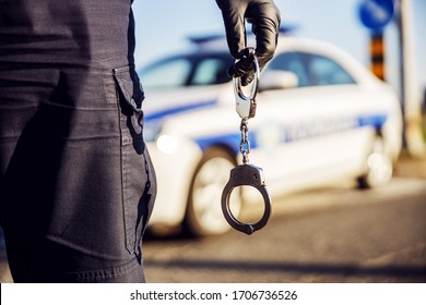 Police Officer Holding Handcuffs, Police Car In Background.