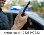 Police officer is holding a walkie talkie near a patrol car with flashing lights