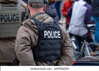 Police officer, in SWAT clothing, standing in a crowded street. The officer is wearing a bulletproof vest with the word police on his back. A group of young people is in the background. 