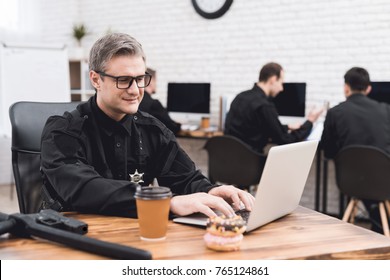 A Police Officer Works For A Laptop In A Police Station. On His Desk Lie Donuts And A Cup Of Coffee. He's In A Good Mood.