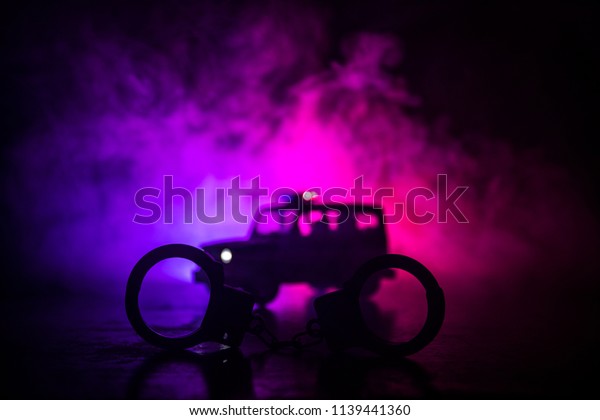 Police raid at night and you are under arrest\
concept. Silhouette of handcuffs with police car on backside. Image\
with the flashing red and blue police lights at foggy background.\
Slider shot