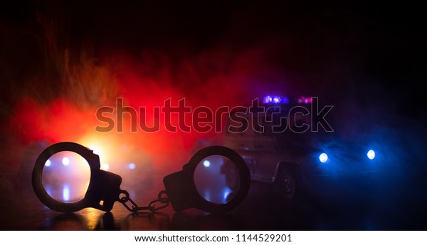Police raid at night and you are under arrest\
concept. Silhouette of handcuffs with police car on backside. Image\
with the flashing red and blue police lights at foggy background.\
Slider shot
