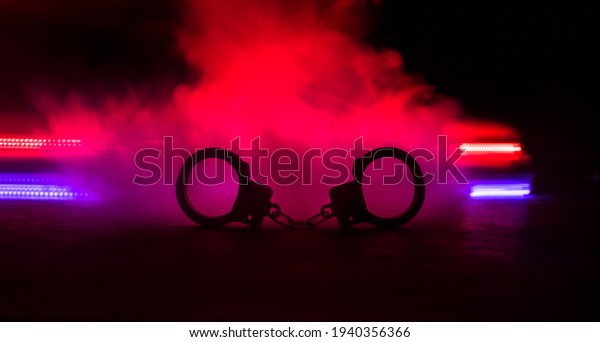 Police raid at night and you are under arrest\
concept. Silhouette of handcuffs with police car on backside. Image\
with the flashing red and blue police lights at foggy background.\
Selective focus
