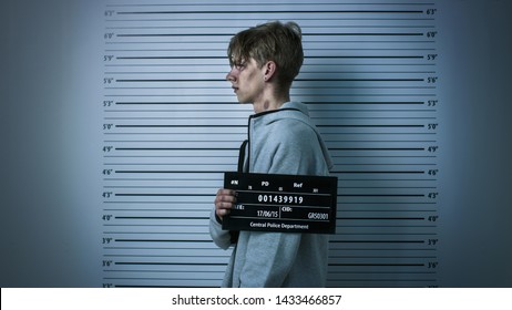 In A Police Station Arrested Drug Addict Teenage Posing For A Side View Mugshot. He Is Heavily Bruised. Height Chart In The Background.