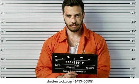 In a Police Station Arrested Man Getting Front-View Mug Shot. He's Wearing Prisoner Orange Jumpsuit and Holds Placard. Height Chart in the Background.