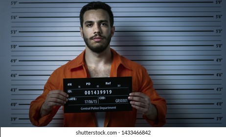 In a Police Station Arrested Man Getting Front-View Mug Shot. He's in a Prisoner Orange Jumpsuit and Holds Placard. Height Chart in the Background.