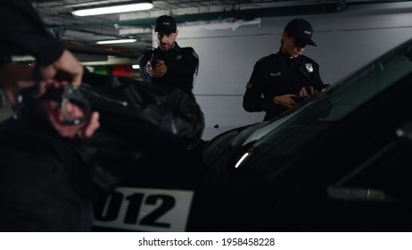 Police team arresting criminal at underground parking. Male cop handcuffing suspect at patrol car. Police officer aiming robber with gun. Policewoman using digital tablet for work