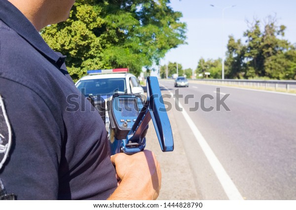 Policeman holding laser\
speed gun near police car on highway background. Selective focus,\
part of body.