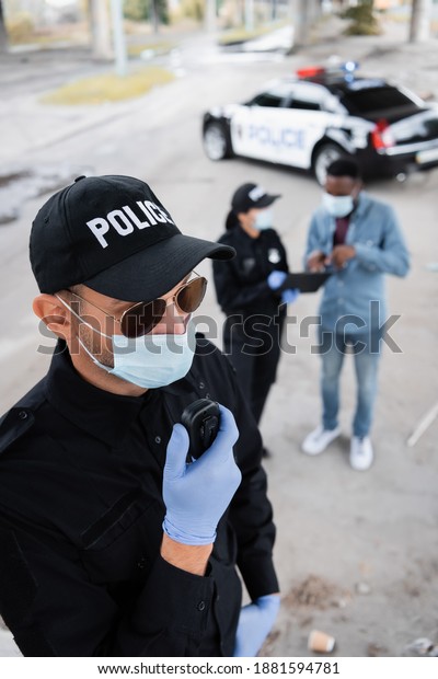 Policeman in latex gloves and\
medical mask using walkie talkie while colleague talking with\
african american victim on blurred background on urban\
street