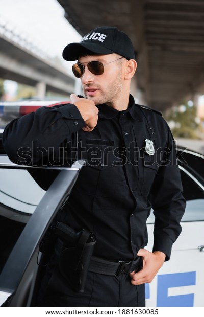 Policeman in sunglasses using walkie talkie near
car on urban street