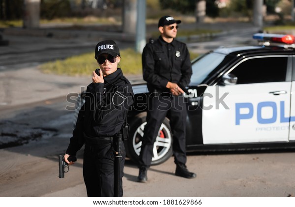 Policewoman with gun\
using walkie talkie near colleague and police car on blurred\
background on urban\
street