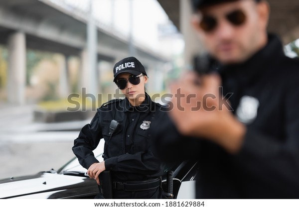 Policewoman in sunglasses taking gun\
near car and colleague on blurred foreground\
outdoors