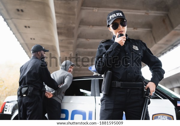 policewoman in\
sunglasses talking on radio set near african american colleague\
arresting offender on blurred\
background