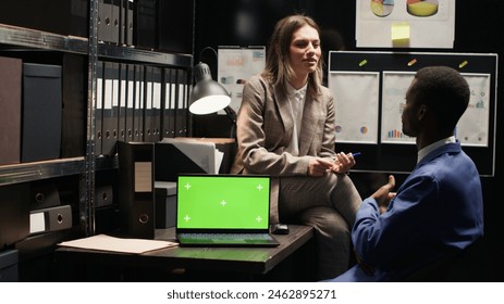 Policewoman talks with black man drinking coffee in office, with laptop displaying greenscreen placed on table. Wireless computer with isolated mockup template on desk while detectives discuss a case. - Powered by Shutterstock