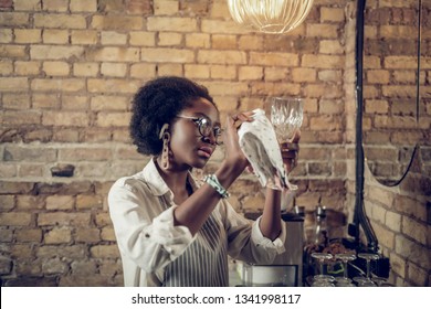 Polishing glasses. Delightful pretty African-American bartender with massive plastic earrings wearing glasses and white shirt with apron polishing wine glass - Powered by Shutterstock