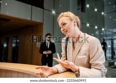 polite blonde woman in suit gesturing while talking at reception desk, personal security service concept, bodyguard in suit standing on blurred background, hotel industry