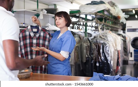 Polite Female Worker Of Dry Cleaner Standing At Reception Counter, Returning Clean Clothes To African American Man 