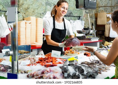 Polite Glad Cheerful Positive Female Fishmonger Standing Behind Counter Of Seafood Store Demonstrating Raw Bonito Steak To Woman Buyer