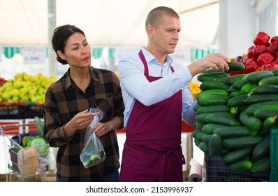 Polite Greengrocery Store Owner Helping Interested Asian Woman In Choosing Fresh Organic Cucumbers