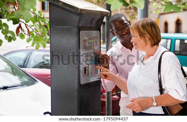 Polite intelligent cheerful positive smiling\
African man helping middle aged woman to buy ticket in parking\
meter on summer city\
street