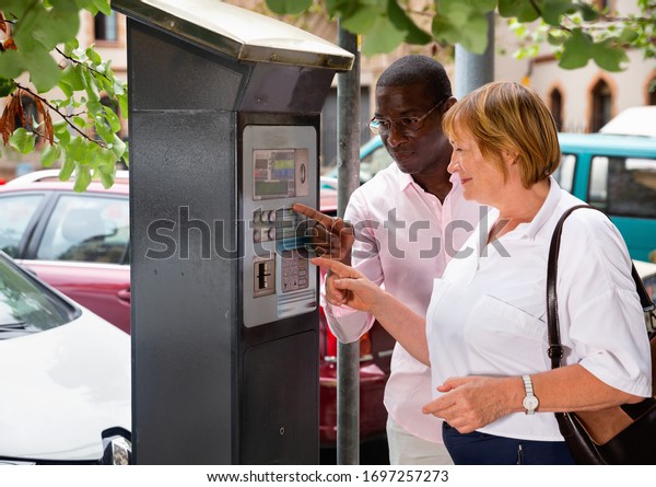 Polite intelligent glad cheerful  smiling African\
man helping middle aged woman to buy ticket in parking meter on\
summer city street