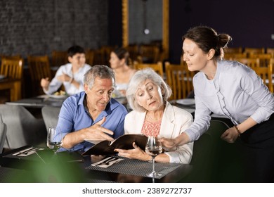 Polite smiling waitress talking to positive elderly couple, man and woman, visiting restaurant for romantic dinner, recommending dishes from menu card - Powered by Shutterstock