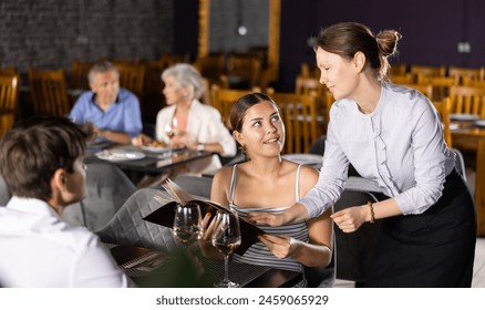 Polite smiling waitress talking to positive young couple visiting restaurant for romantic dinner, recommending dishes from menu card - Powered by Shutterstock