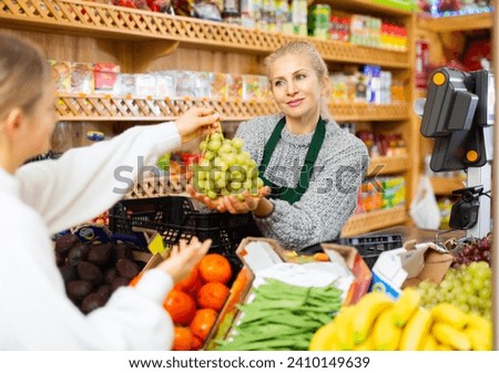 Polite woman seller standing near scales behind counter, taking bunch of ripe grapes from female shopper for weighing