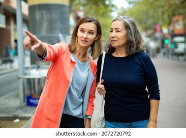 Polite Young Woman Pointing Way To Aged Female Tourist On City Street On Warm Autumn Day.