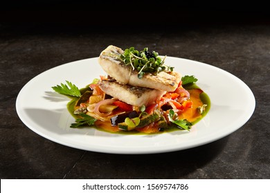 Pollock Fish Fillet With Vegetables On White Plate. Gourmet Delicious Food. Baked Fish Piece With Sweet And Sour Sauce And Herbs Side View. Seafood With Steamed Veggies Meal On Black Table