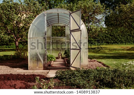 Polycarbonate greenhouse in the garden for growing your own fresh food with doors open and a watering can in the centre in late afternoon light. Compact DIY solution for gardens.