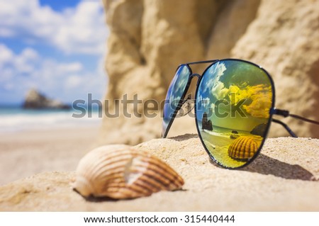 Poly-color sunglasses, reflecting the ocean and a shell on a sandy, tropical beach